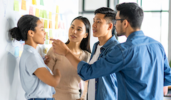 Students stand at a white board discussing items on post-it notes.